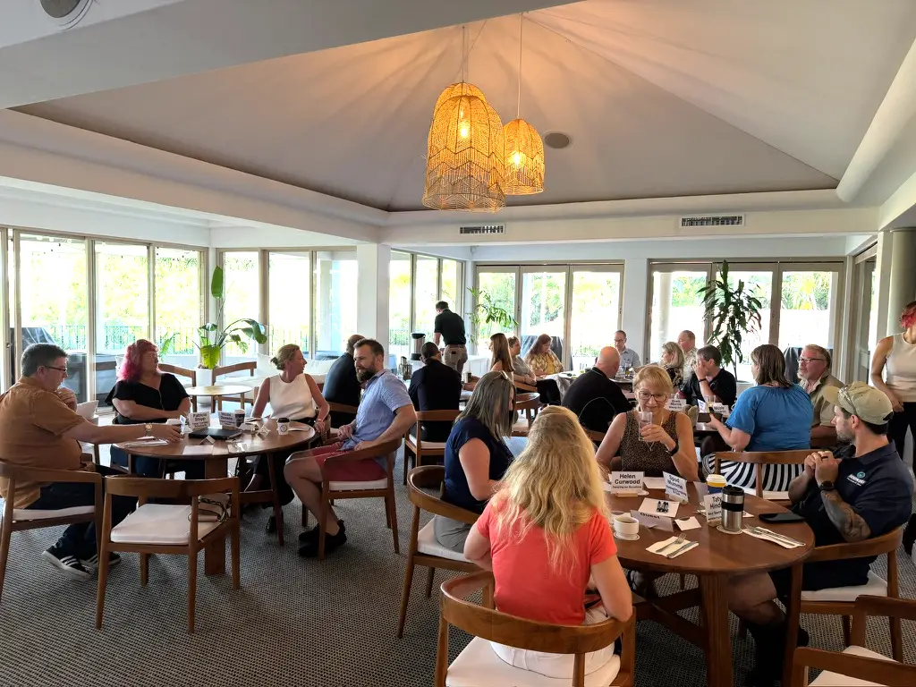 Noosa Business Group members chatting at round timber tables with name cards, in a bright clubhouse room with large windows and woven pendant lights.