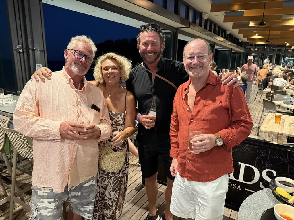 Four Noosa Business Group members smiling with drinks on a timber deck at an outdoor venue in the evening, with diners behind them.