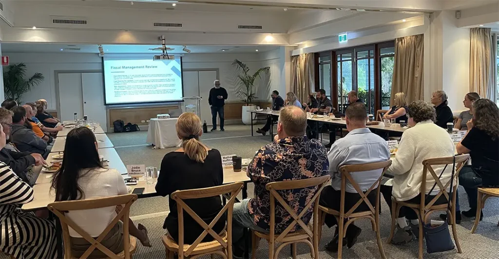 A guest speaker presenting a “Fiscal Management Review” slide to Noosa Business Group members seated along long breakfast tables.